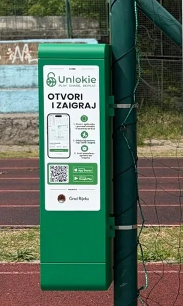 Zeleni Unlokie ormarić stoji uz stup pokraj atletske staze, na njemu upute i QR kod